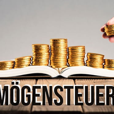 Bild von einem Holztisch auf dem ein geöffnetes Buch liegt. Auf diesem Buch liegen Geld-Münzen und eine Hand legt weitere dort drauf. Unten kann man den Schriftzug Vermögensteuer und einer spanischen Flagge daneben.