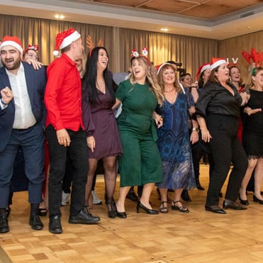 30 neue Mitarbeiter der PlattesGroup singen und tanzen auf der Weihnachtsfeier der Firma. Sie tragen alle festliche Kleidung und haben gute Laune